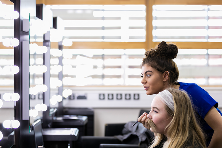 Two students looking in mirror with one applying makeup to the other